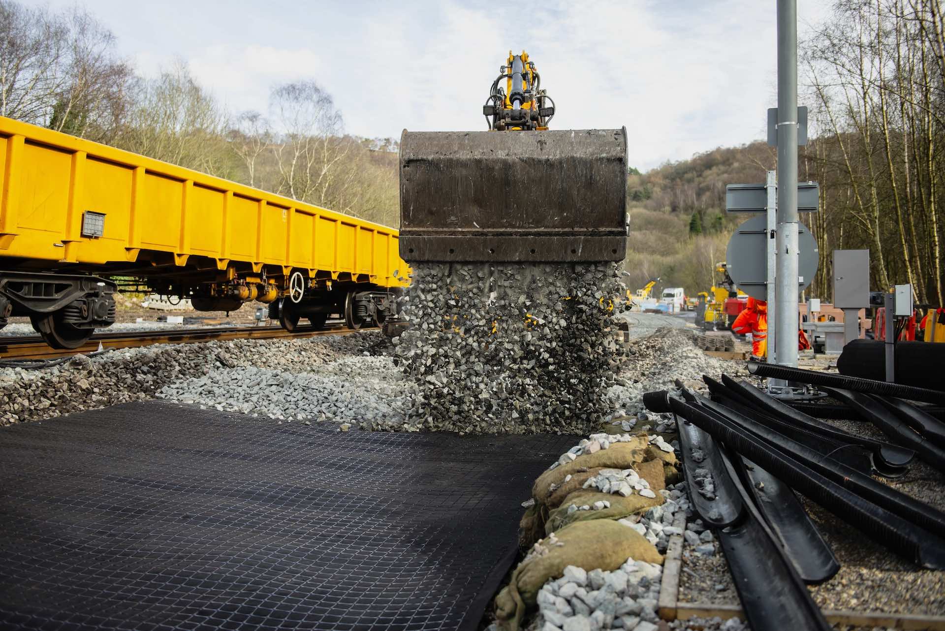 Construction site showing a large excavator bucket pouring gravel onto the ground, near railway tracks. A yellow train car is on the left, and workers in orange safety gear are visible in the background. Trees and hills are in the distance.