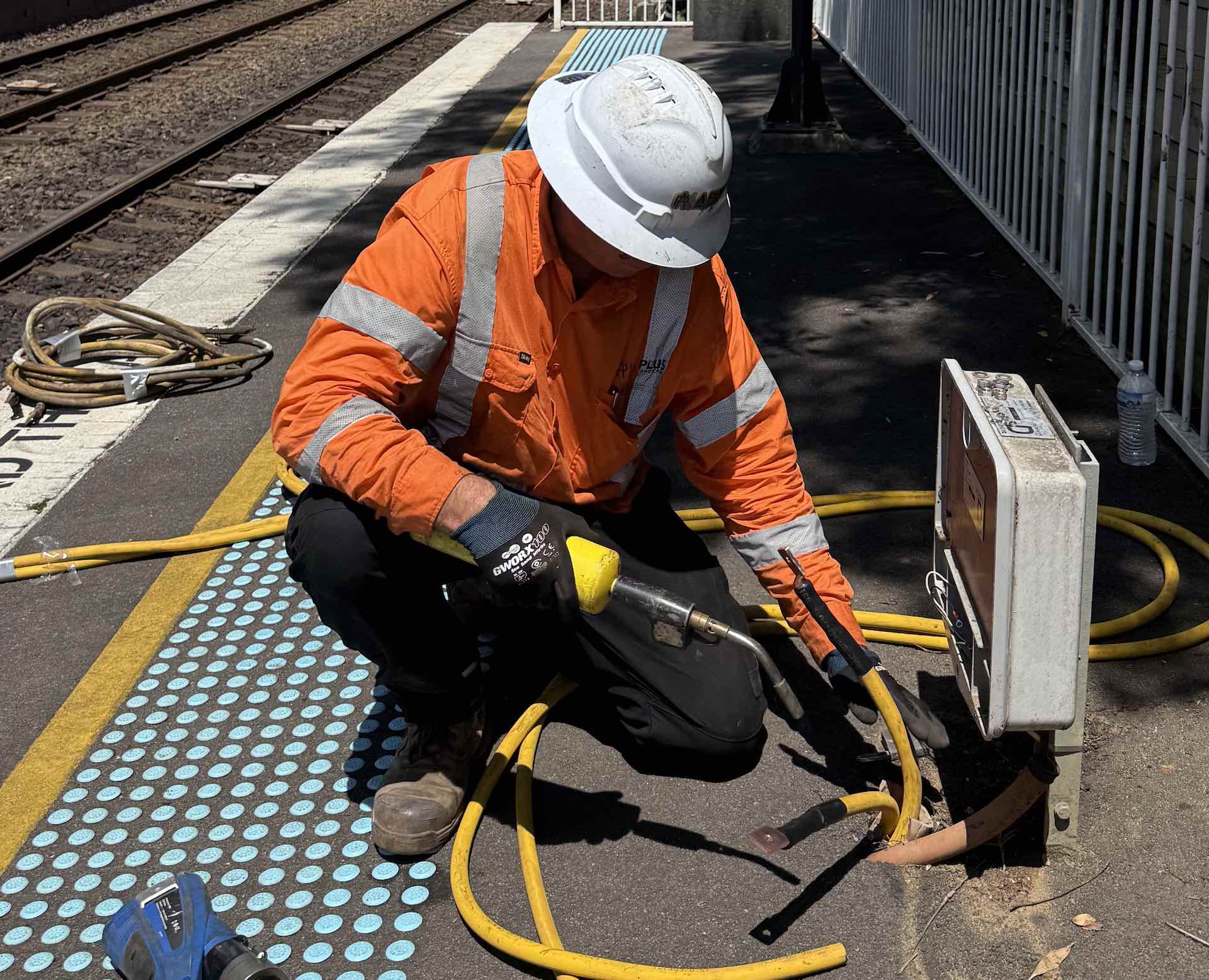 A worker in an orange high-visibility jacket and white hard hat uses a power tool to repair equipment on a train station platform. Nearby are hoses and tools. Safety gloves are worn. Tracks and a railing are visible in the background.