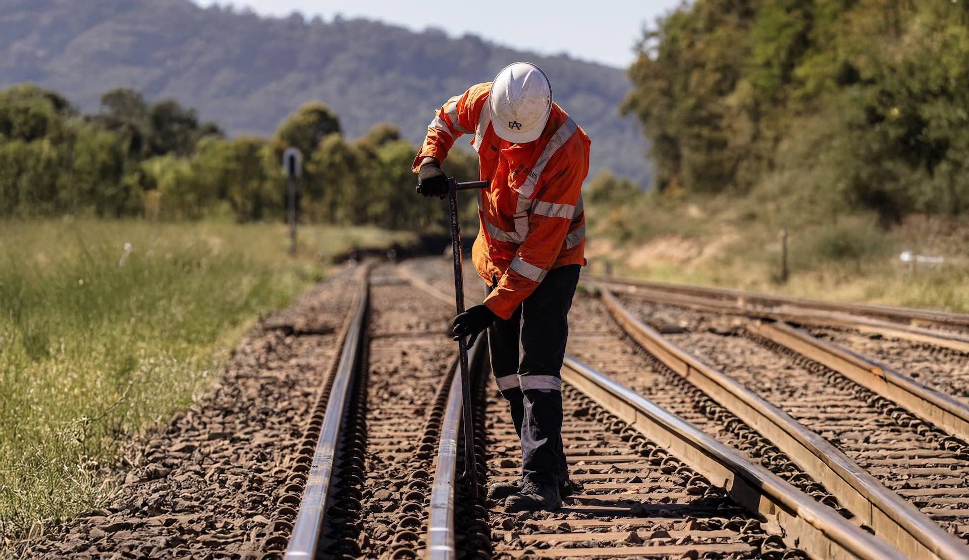 A construction worker in an orange safety jacket and white helmet is using a tool to adjust a railway track. The scene is set outdoors with trees and hills in the background under a clear sky.