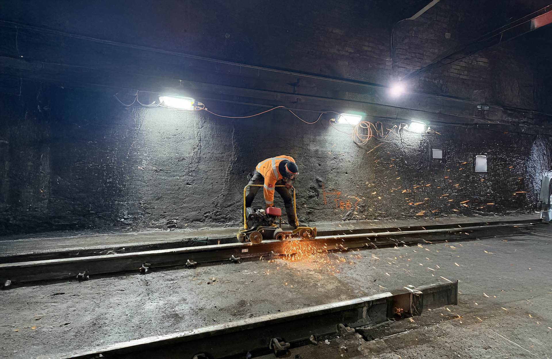 Worker in an orange jacket grinding metal on railway tracks in a dimly lit industrial setting. Sparks fly around as the grinder makes contact with the metal. Overhead lights illuminate the scene.