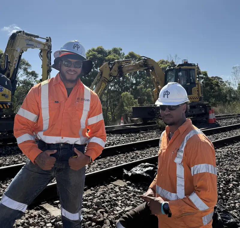 Two construction workers wearing orange safety jackets and hard hats stand on a railway track. Behind them, two excavators and orange cones are visible. The sky is clear, and trees are in the background.