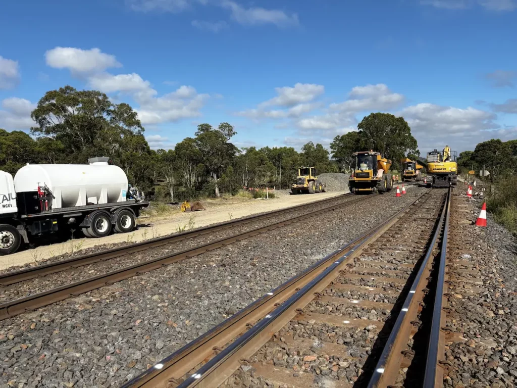 Railroad tracks under construction with heavy machinery and equipment on site, including a tanker and bulldozers. Orange cones line the area. Trees and a cloudy sky are in the background.