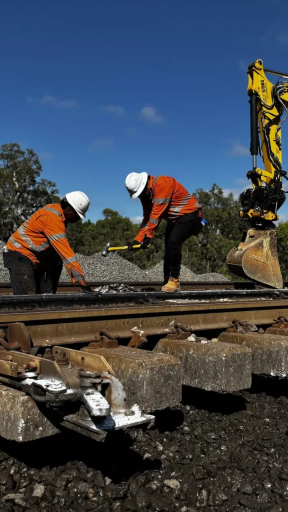 Two construction workers in orange safety gear and white helmets work on a railway track. One holds a tool, while an excavator is nearby. The sky is clear, and there are trees in the background.