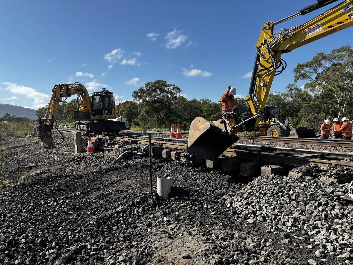 Two excavators and workers are repairing railway tracks in a rural area with trees and hills in the background. One excavator lifts a track section while workers in orange safety gear assist. Blue sky and scattered clouds above.