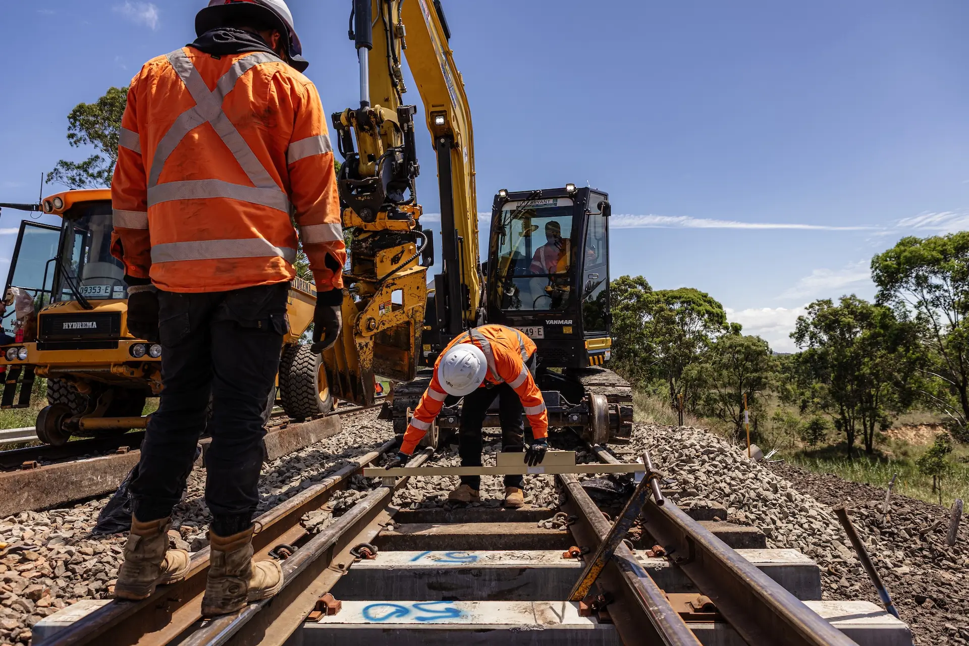 Two construction workers in orange safety gear work on a railway track. One operates a machinery vehicle, while the other adjusts the tracks. Trees and a clear sky are in the background.